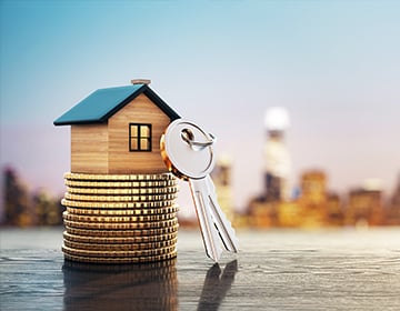 miniature wooden house coin stacks with keys against blurred city skyline